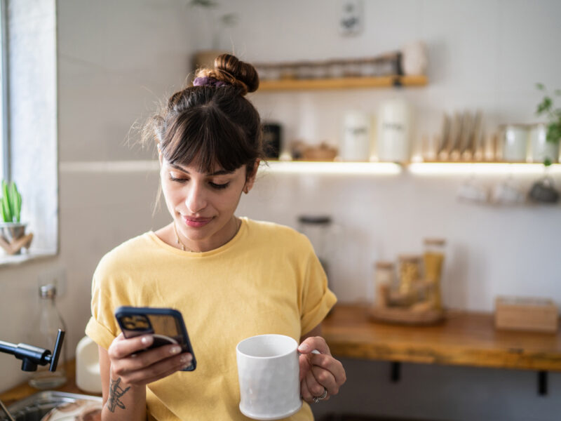 Young woman wearing a yellow shirt looking at phone and texting in kitchen while holding a mug