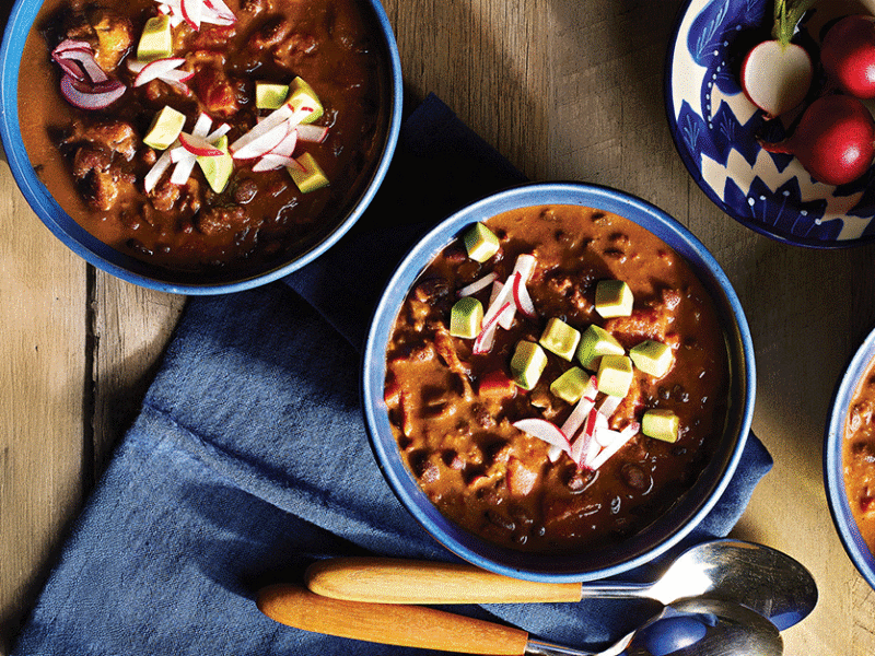 Three bowls of Mole Chili on a wooden table, with a blue napkin, wooden-handled spoons, and blue cermamic bowls
