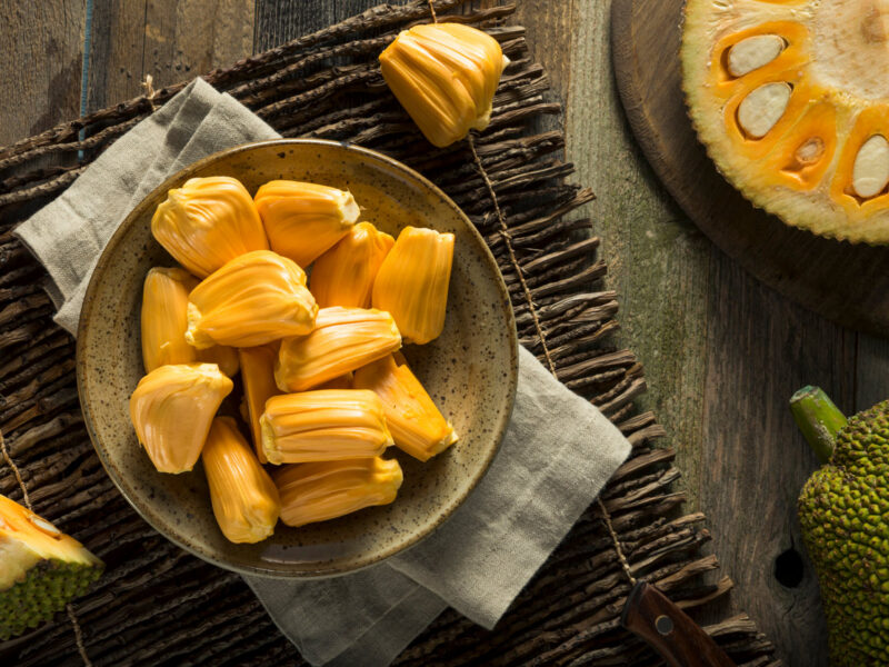 Jackfruit pods in a bowl on a wooden table next to a halved jackfruit with pod segments visible