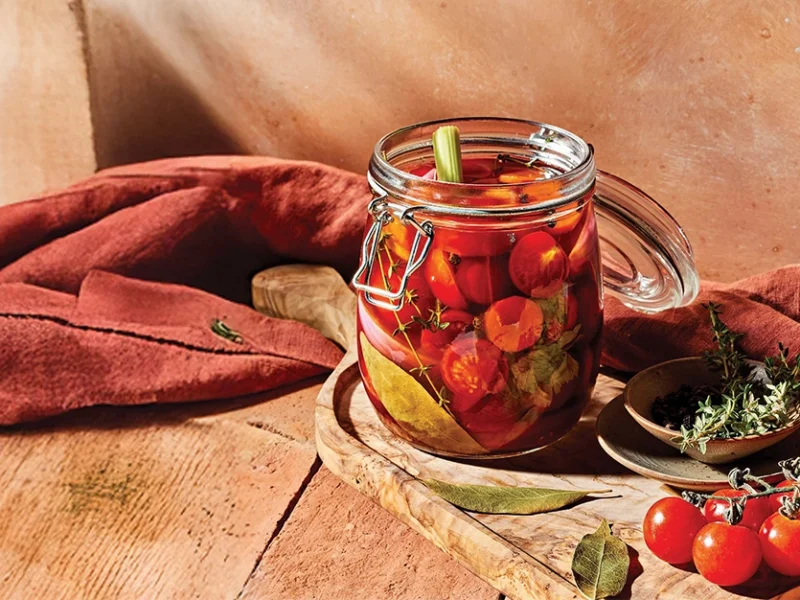 A jar of Quick-Pickled Cherry Tomatoes on a wooden counter, with a dark red kitchen towel behind and fresh cherry tomatoes to the side