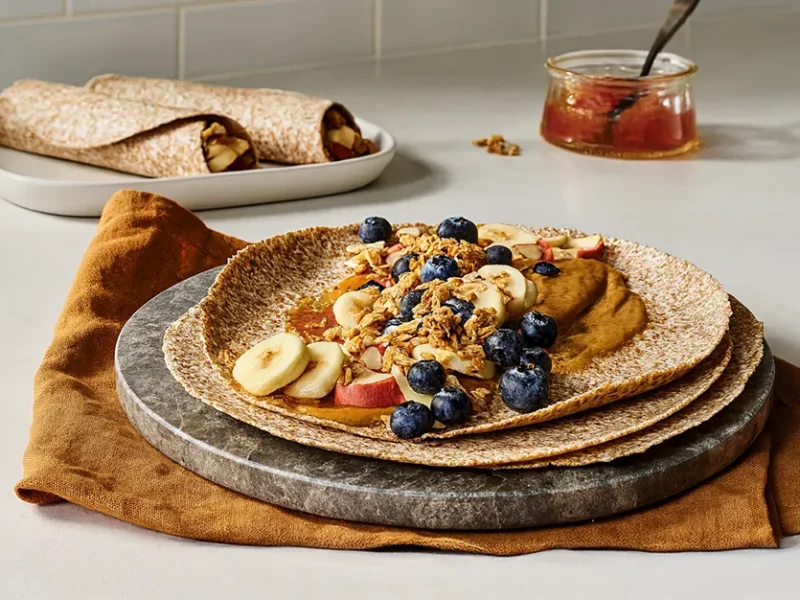 A whole grain tortilla topped with P B and J and topped with banana slices, berries, and granola