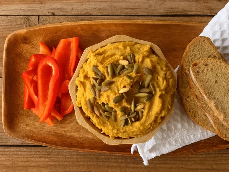 Pumpkin Hummus on a wooden serving platter with sliced red bell peppers and slices of crusty bread