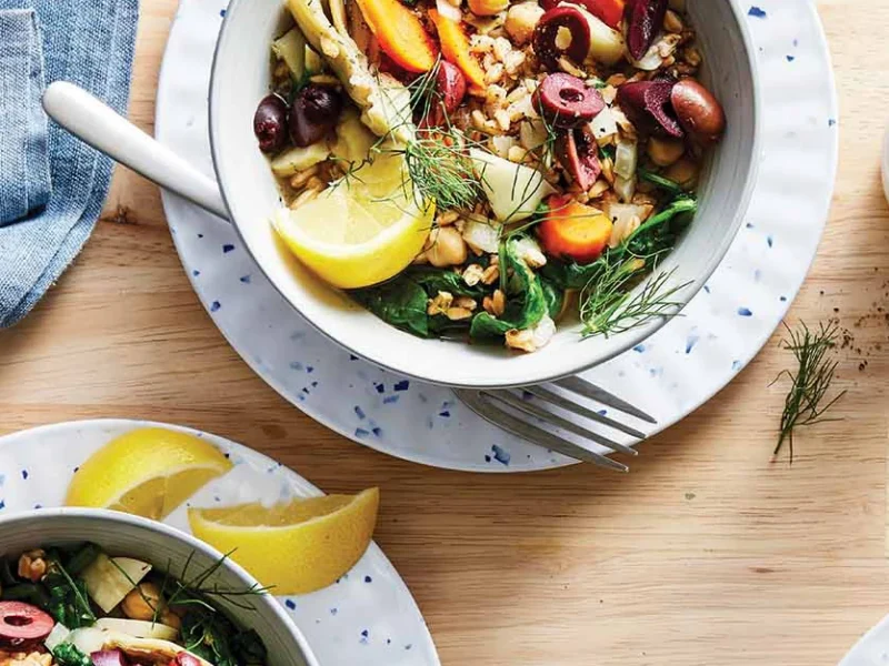 Two white serving bowls on white plates with lemon wedges on the side. Each bowl is full of a colorful mix of Greek-Style Farro with Chickpeas and Artichokes