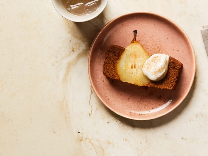 A slice of Gingerbread Loaf with Poached Pears on a pale pink plate, topped with a dollop of Aquafaba Whipped Cream.