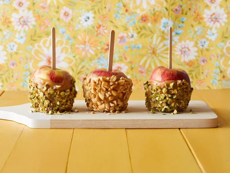 three vegan caramel apples dipped in nuts on a yellow background