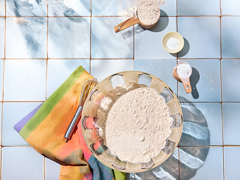 Ingredients for a healthy homemade pancake and waffle mix, with the mix in a large mixing bowl on a light blue tile surface, with a colorful kitchen towel and whisk beside it