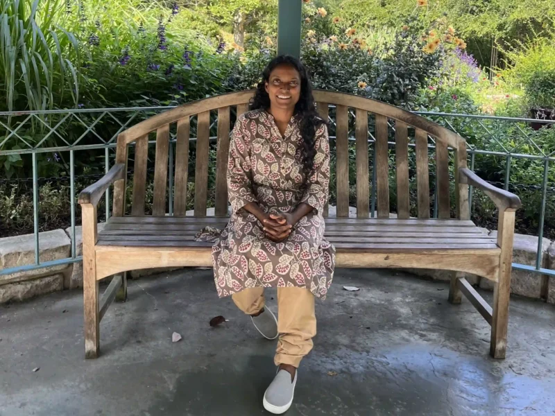 Nurse practitioner Maxine Charles sitting on a bench in a gazebo on a sunny day, wearing a leaf-print dress and khaki pants, smiling, after lowering her A1c on a plant-based diet and reversing prediabetes