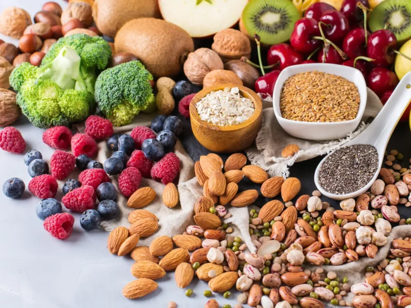 Assorted whole plant foods rich in antioxidants, including broccoli florets, berries (raspberries, blueberries, cherries), kiwi, apples, oats, flaxseeds, chia seeds, almonds, walnuts, and a variety of dried and cooked beans, arranged on a light surface.