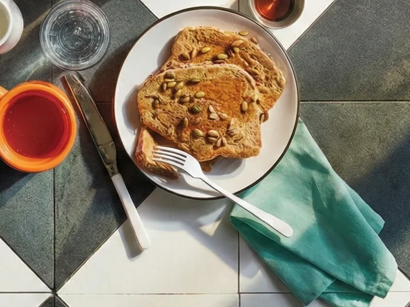 Two slices of Whole Grain French Toast on a breakfast setting with a small cup of maple syrup and a cup of tea and glass of water