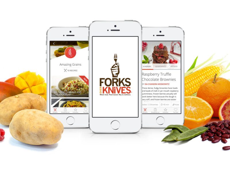 Three mobile phones showing the Forks Over Knives app, on a white background next to fresh fruits and vegetables
