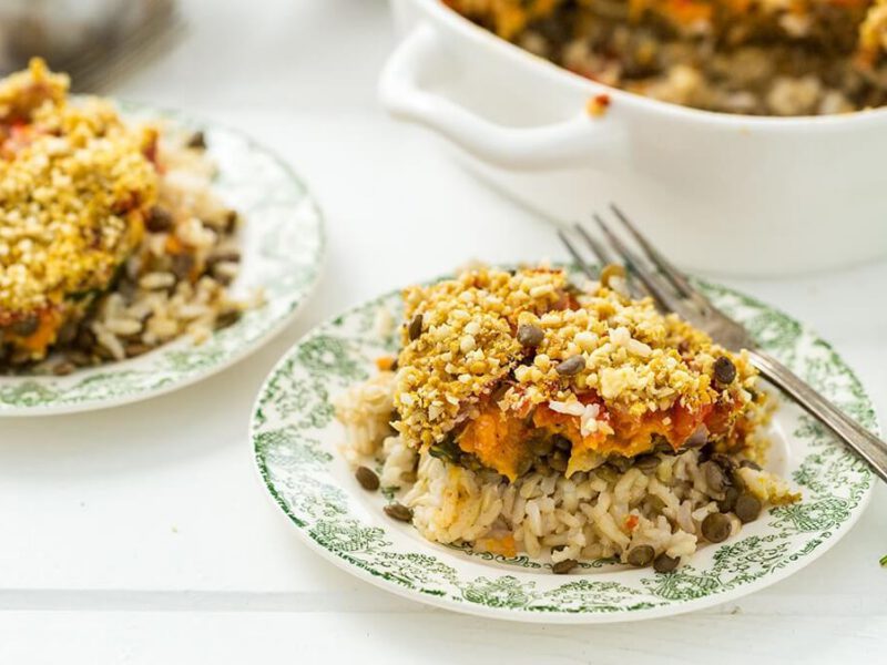 Two plates of Vegan Rice Casserole with Lentils, with a fork on the side. In the background is a large white casserole dish filled with the rice and lentil mix.