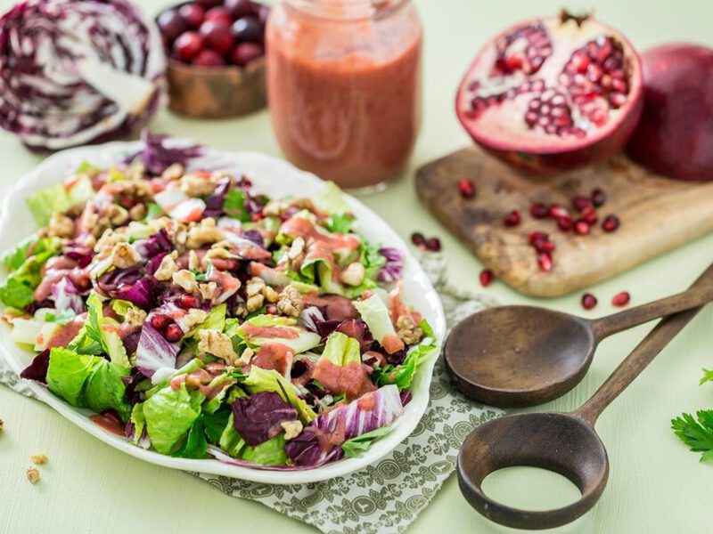 A large summery spread on a pale green table cloth, which includes a platter of Radicchio Salad with Cranberry Orange Dressing, wooden servings spoons, fresh pomegranates, and half a radicchio lettuce