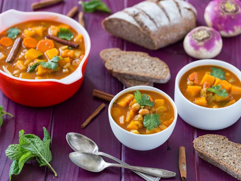 A large pot, and two smaller bowls of Vegetable Autumn Stew on a purple wooden table with chunks of whole grain bread and a couple of spoons