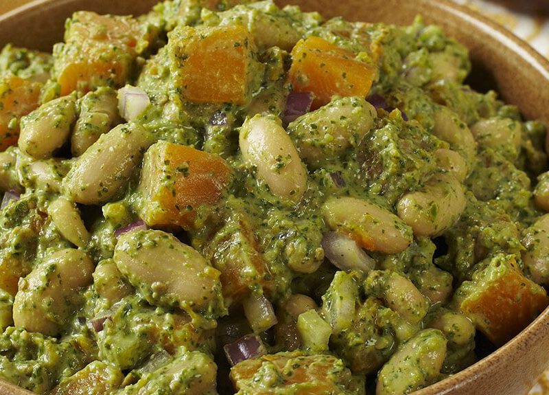 A brown ceramic bowl of Winter White Bean Salad with Spicy Cilantro Pesto. In the background is a wooden serving spoon and a smaller wooden bowl of pesto