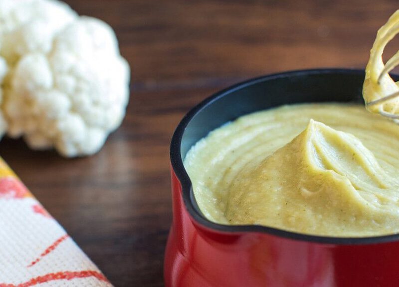 Cauliflower Bechamel in a small red pot, with a hand whisk held above the bowl and a fresh head of cauliflower in the background