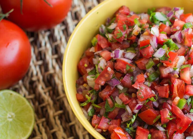 Fresh homemade tomato salsa in an yellow bowl beside fresh tomatoes, limes, and herbs
