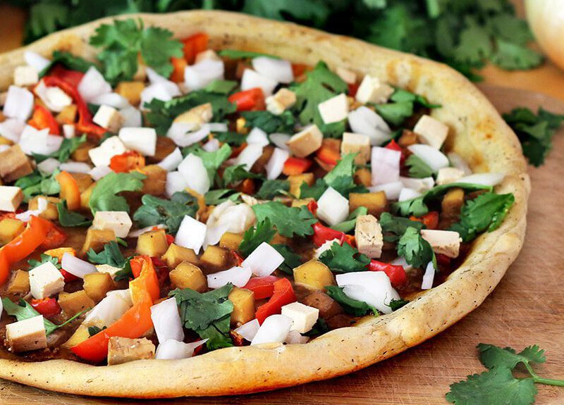 Vegan pizza topped with colorful chopped veggies atop a wooden chopping board with an onion and sprigs of parsley in the background