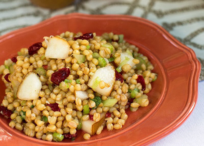 A terracotta colored bowl of wheat berry salad with pear and cranberries dotted throughout