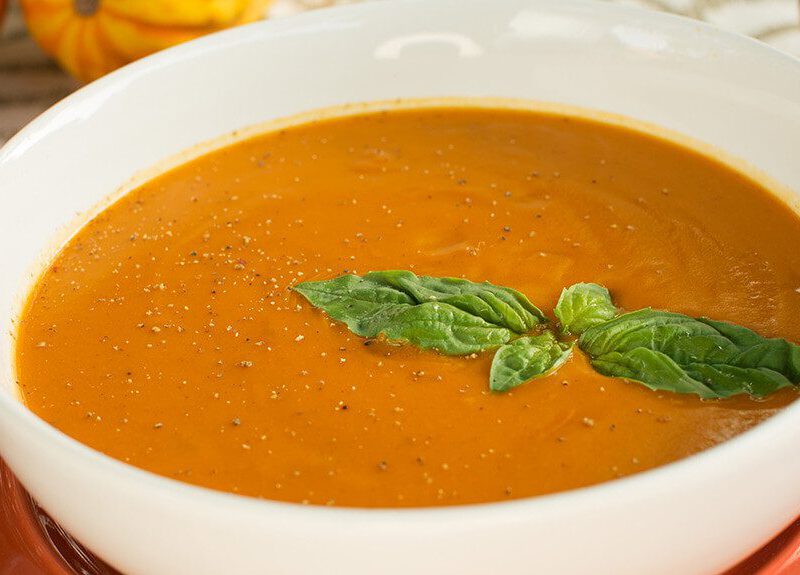 A white bowl of Sweet-Potato-Bisque on a red plate, with a sprig of fresh basil on top
