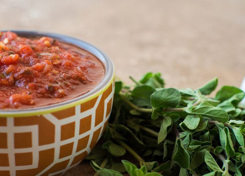 Tomato Sauce in a decorative brown ceramic bowl next to a bunch of fresh herbs and a head of garlic.