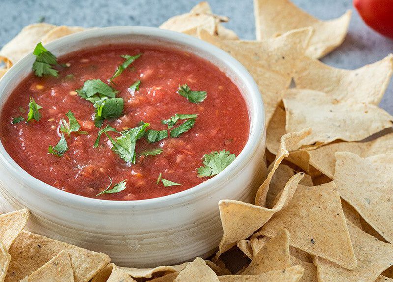 Roasted Tomato Salsa with baked chips next to fresh tomatoes and a few cilantro leaves