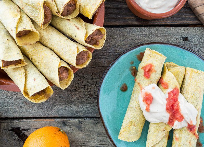 A serving bowl full of Orange Black Bean Taquitos, with three taquitos on a blue serving plate, topped with salsa and tofu cream