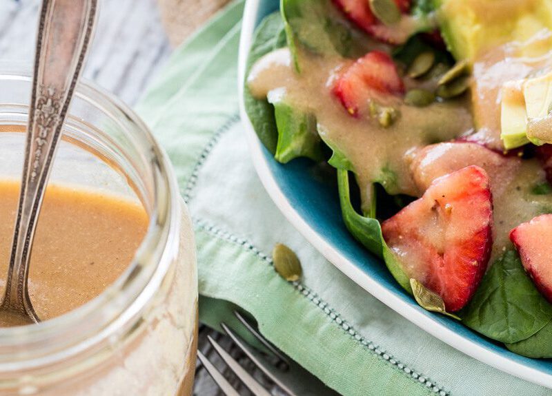 A mason jar of "Magical" Applesauce Vinaigrette next to a salad with fresh strawberries, spinach, and avocado, drizzled with the vinaigrette