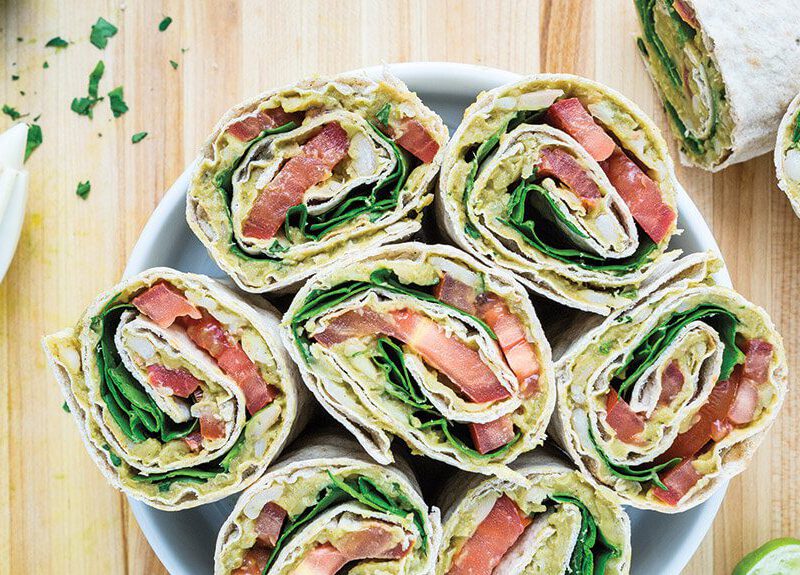 Four Avocado & White Bean Salad Wraps cut in half with the cut side face up, in a white bowl with a a hand-held lemon squeezer to the side