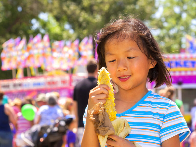 A young girl at a carnival eating an ear of corn