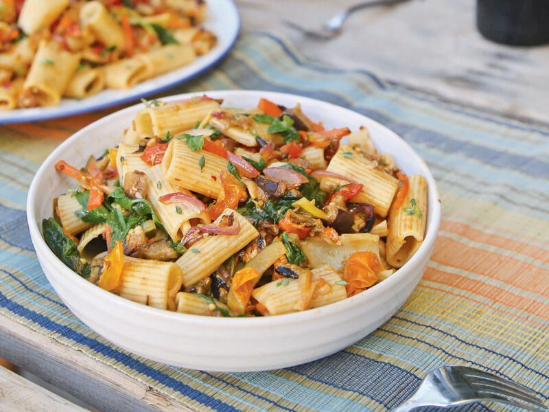 A bowl of roasted veggie pasta and a fork on a summery table mat, with a larger bowl of the mix in the background