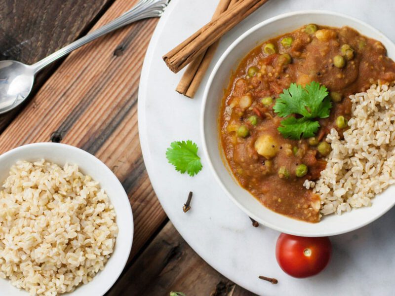 Aloo Mattar in a white bowl with brown rice on a marble chopping board with cinnamon stick and fresh tomatoes in the background