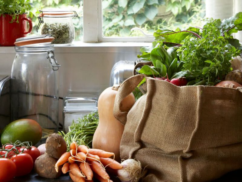 A cloth bag filled with fresh veggies on the kitchen counter next jars, a blender, more veggies