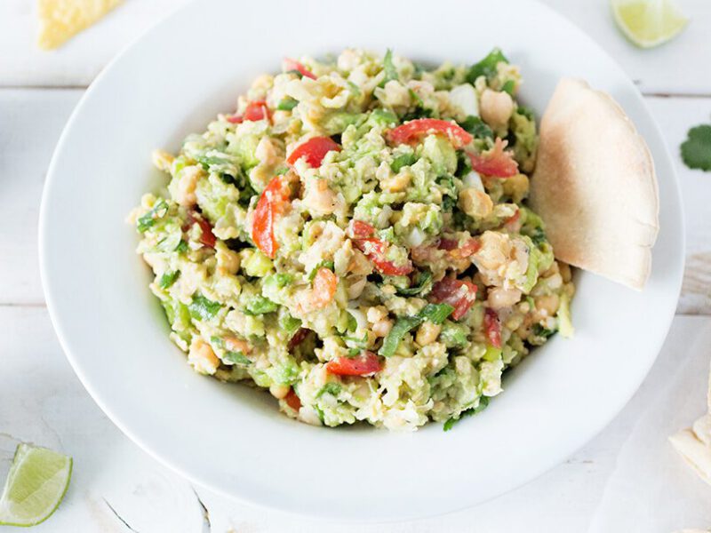 Smashed Chickpea Avocado Dip in a white bowl on a white background with pita bread squares and a whole avocado cut in half off to the side