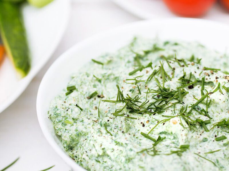 A small bowl of Spinach Ranch Dip garnished with fresh herbs next to a plate of carrot and cucumber sticks