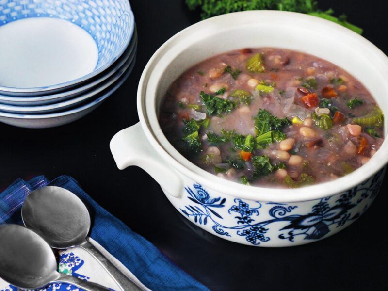A large white pot of Navy Bean Soup next to a stack of blue and white bowls and a couple of spoons