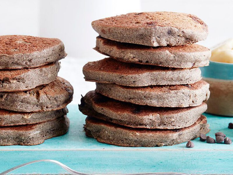 Two large stacks of Chocolate Chip Coconut Pancakes next to a bowl of sliced banana and a small bowl of vegan chocolate chips