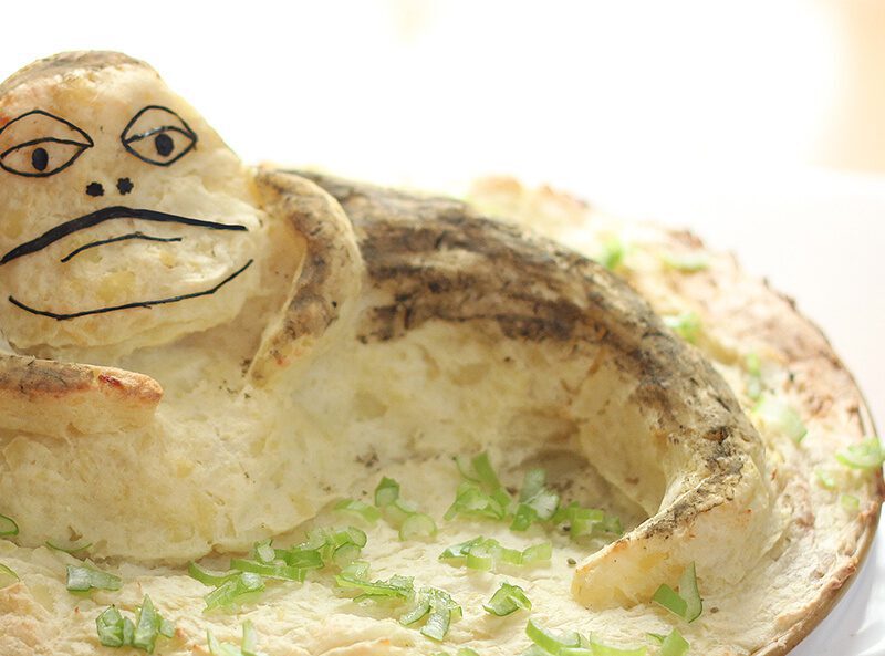 A white dish of shepherd's pie topped with mashed potatoes. The mashed potatoes are piled so they look like a person and have a face draw on them.