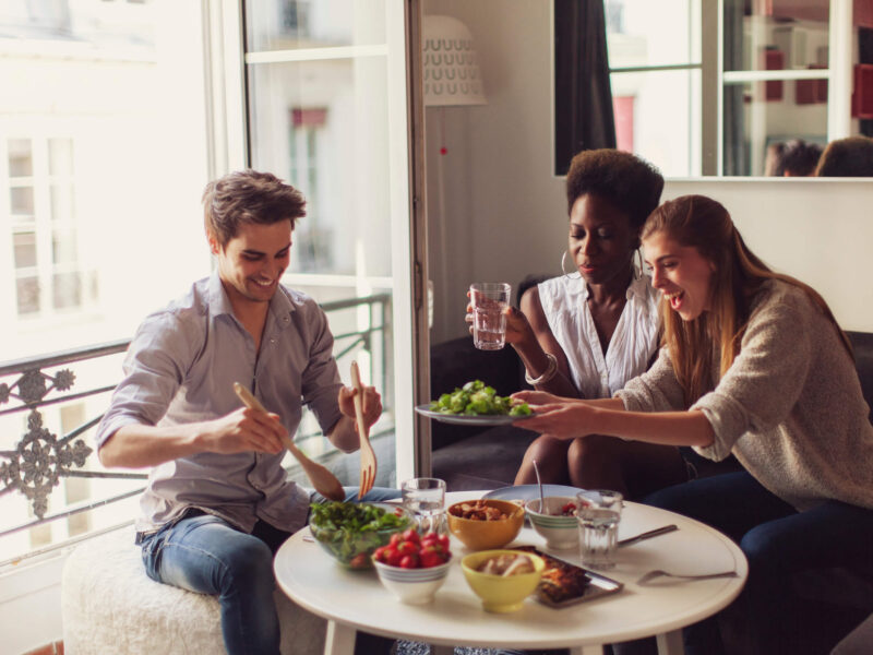 Three young adults laughing and sitting around a small table, sharing healthy plant-based food