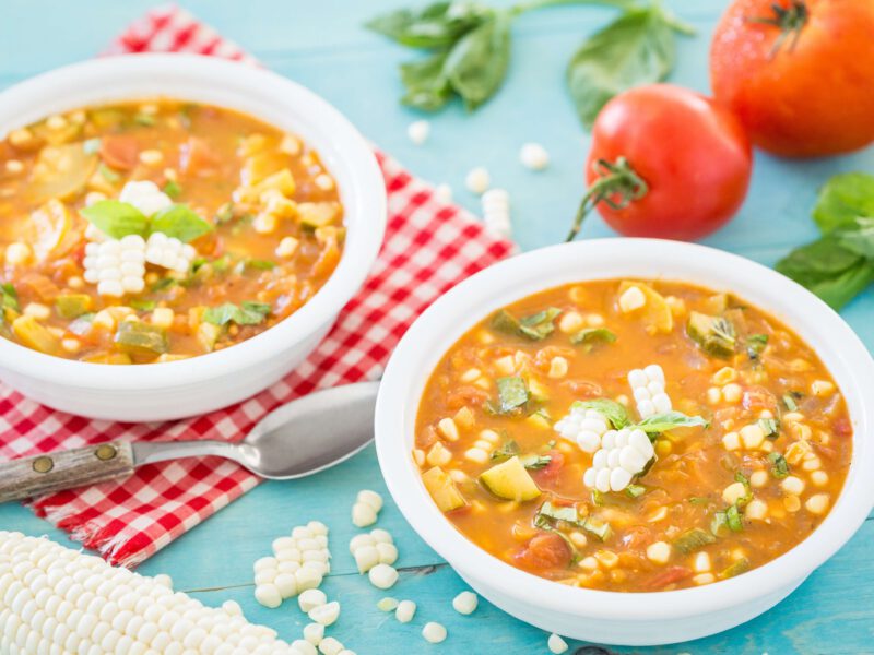 Two bowls of Summer Vegetable Soup garnished with fresh basil and planks of corn, on a red and white checked napkin and blue table. Fresh tomatoes, corn, and basil are in the background.