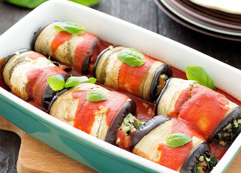 A casserole dish atop a wooden chopping board, filling with six Eggplant Rollatini drizzled in a tomato sauce and garnished with a leaf of fresh basil