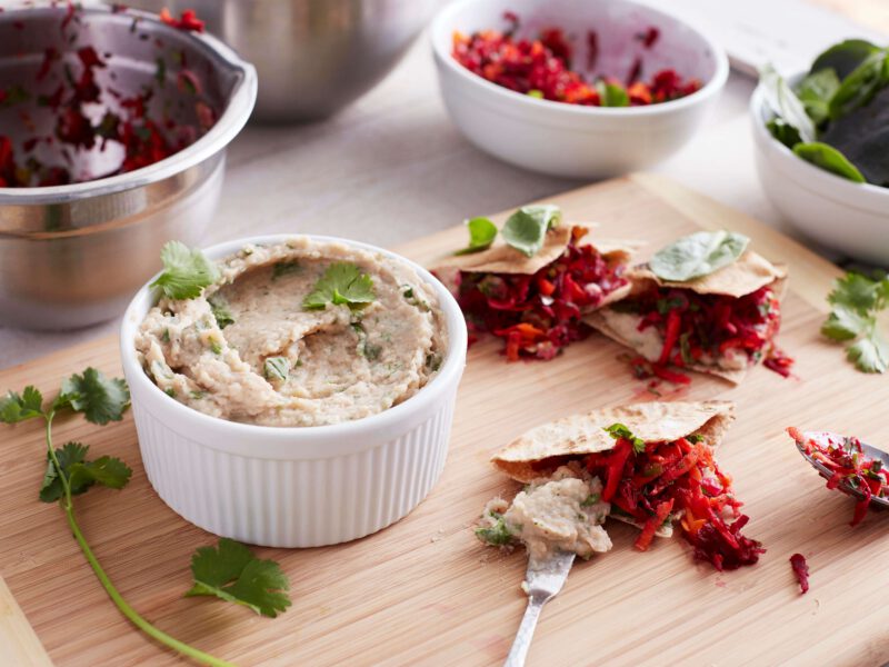 A ramekin of white navy bean hummus sprinkled with fresh herbs is on a cutting board beside whole wheat pitas and fresh veggies