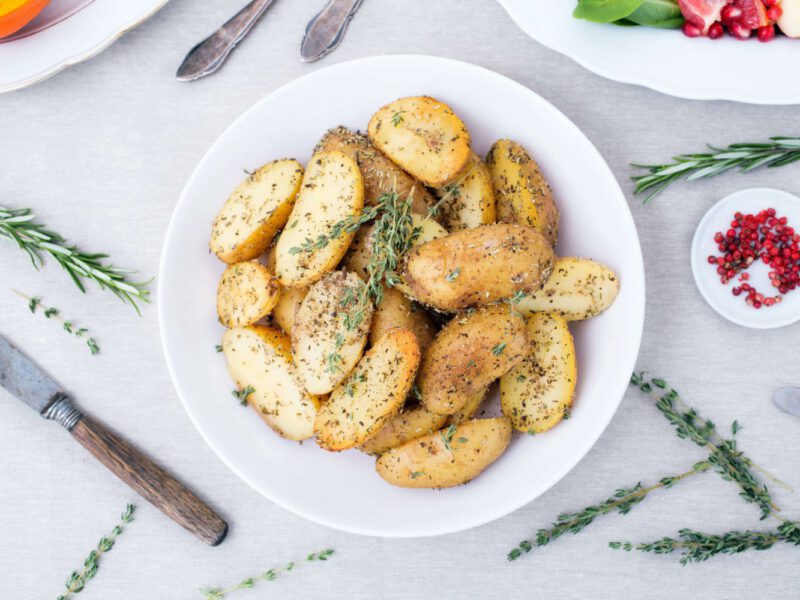 Small, herb-coated, cooked potatoes cut in half presented on a white plate with a sprig of rosemary on top. The plate sits on a marble table top with more rosemary and a small bowl of pomegranate arils beside it.