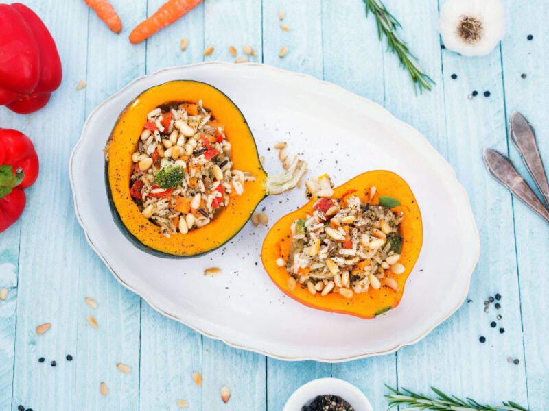 A top down view of two halves of Roasted Stuffed Winter Squash on a white platter, with fresh red bell pepper, carrots and sprigs of rosemary surrounding the platter. The background is a pale blue washed-wood surface.