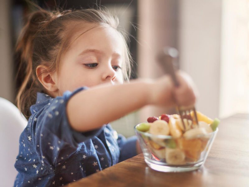 A two-year old girl at the table eating fruit salad with a fork