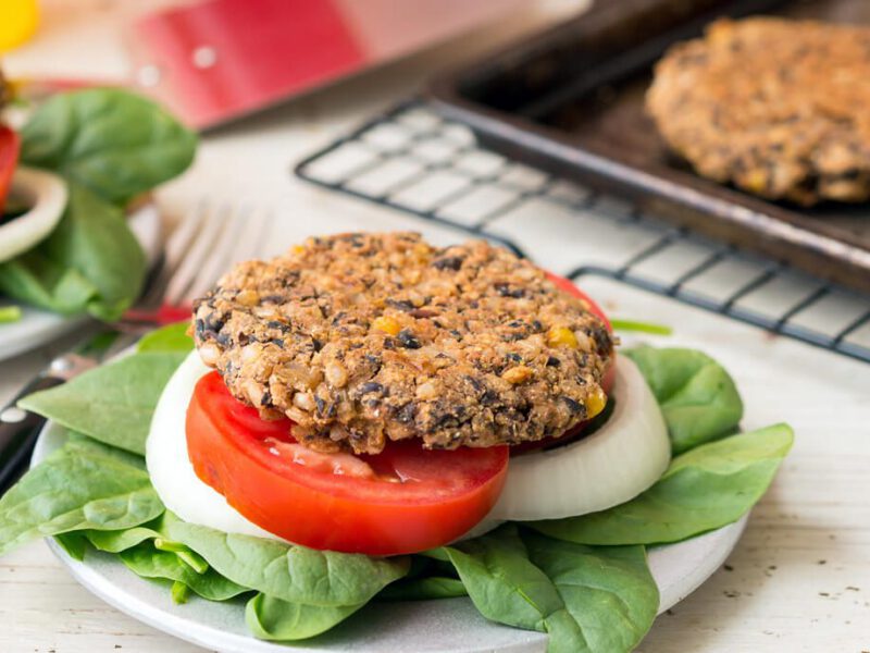 A black bean burger on a plate with fresh greens, a slice of tomato, and a slice of onion
