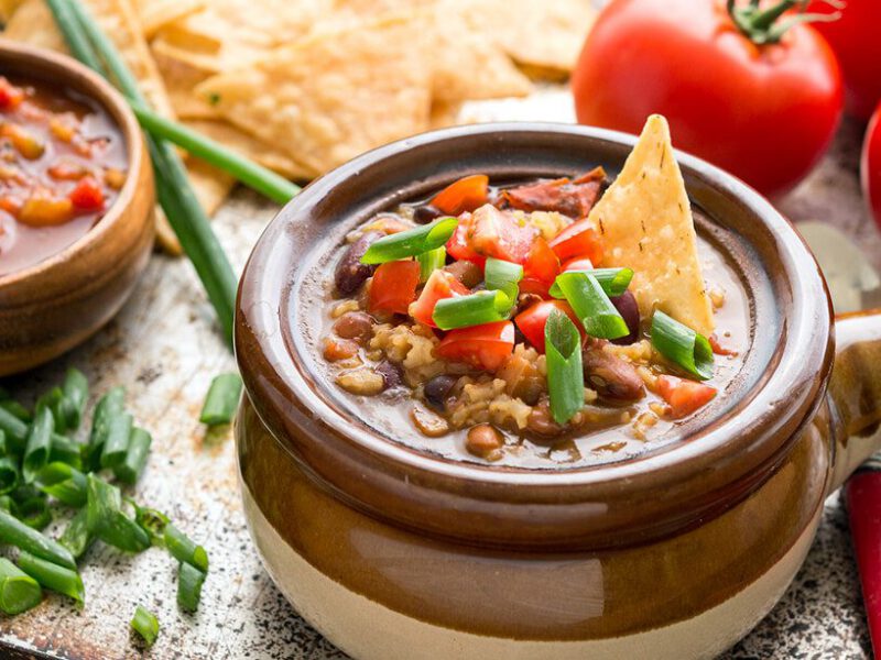 Mexican Rice Soup in a brown soup bowl with a handle on a baking tray along with fresh tomatoes, baked corn chips, salsa, and chopped green onion