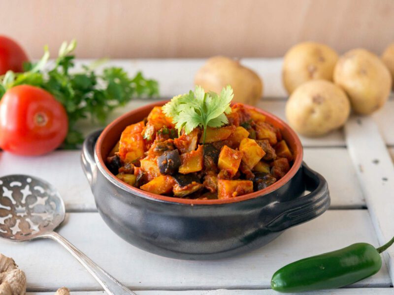 A soup with handless, filled with a hearty stew: Aloo Baingan (Potato and Eggplant). in the background are raw potatoes, tomatoes, parsley, and chili