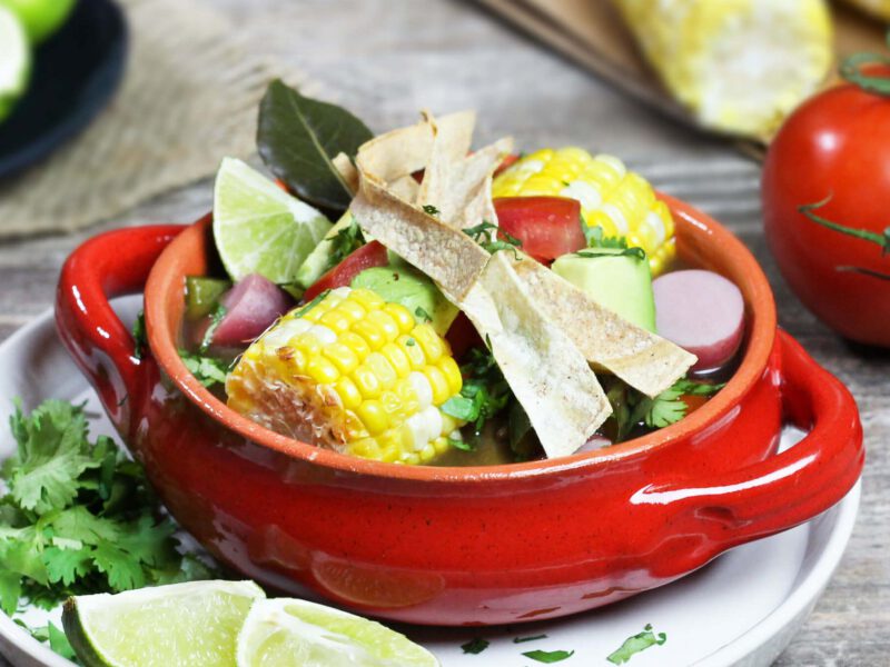Mexican Lime Soup in a white two-handled soup bowl on a plate with lime wedges, fresh cilantro, and with fresh strawberries and corn in the background