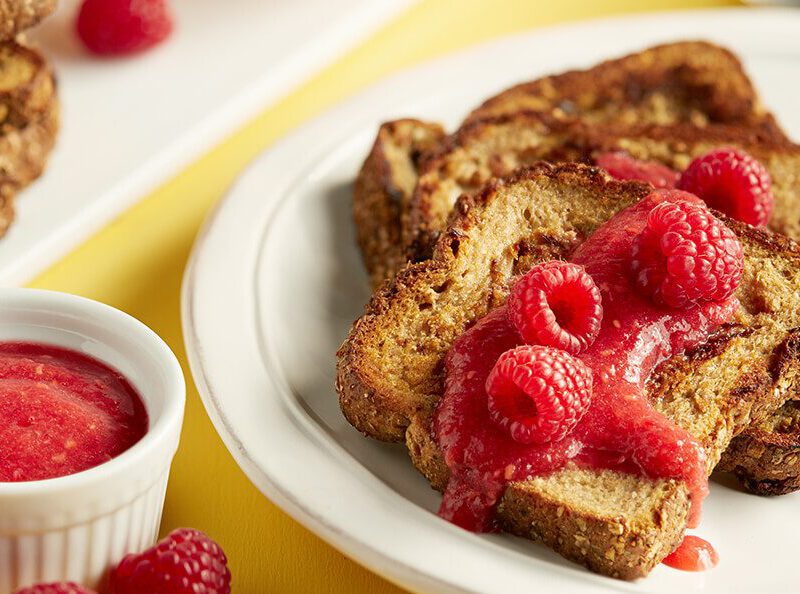 On a bright yellow table is a white plate with vegan Orange French Toast, fresh raspberries, orange slices, and a raspberry compote. Next to the plate is a small white ramekin of extra compote.