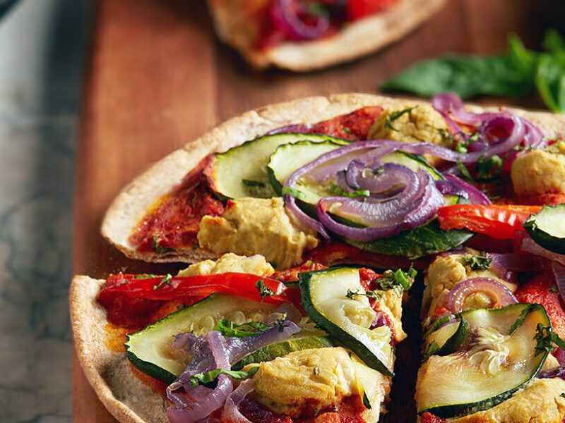 Cheesy Vegan Veggie Pizzette cut into quarters on a wooden chopping board, with other pizzettes in the background